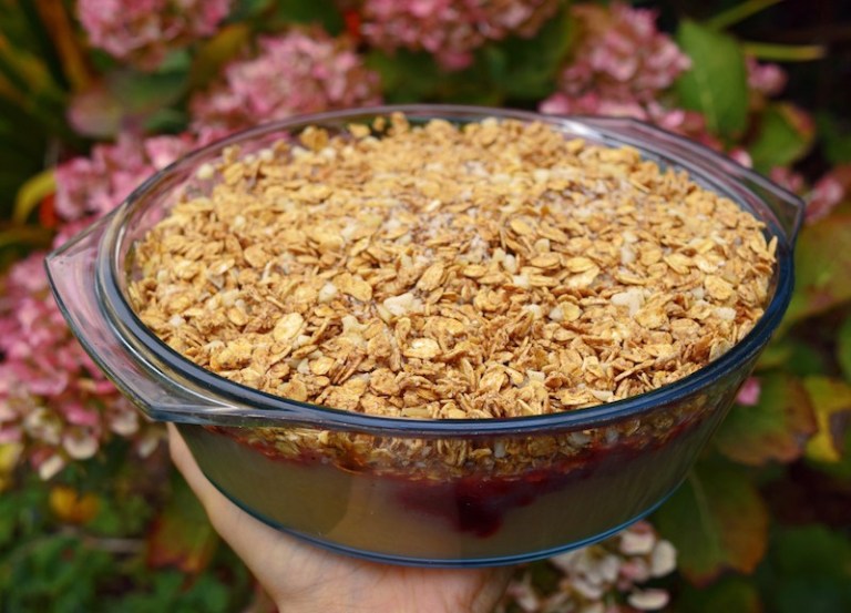 An image of me holding a dish of this apple crumble recipe, with pink flowers in the background. 